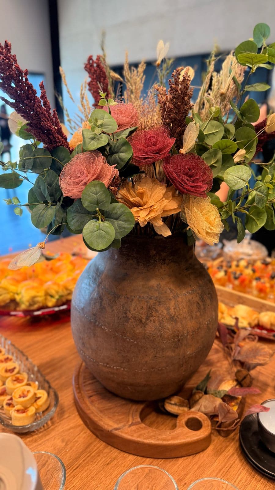 Arranjo floral rústico em vaso de cerâmica com detalhe de salgados ao fundo