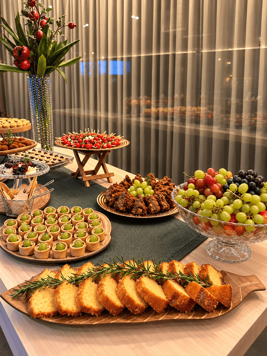 Mesa com bolo de fubá fatiado, brownies, uvas e taça de frutas em evento corporativo
